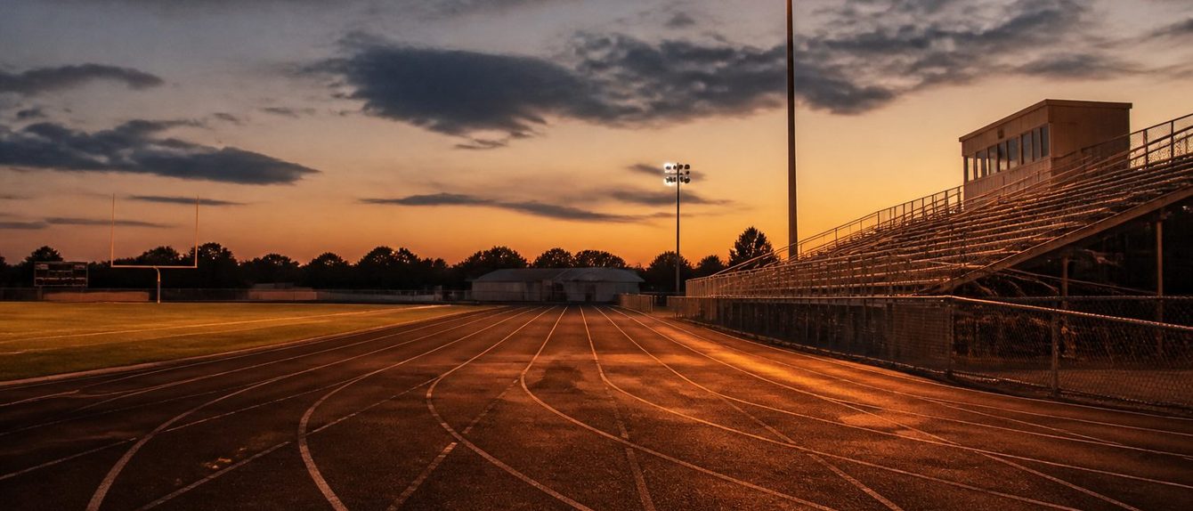 Empty stadium track at dusk with one light tower on above the bleachers